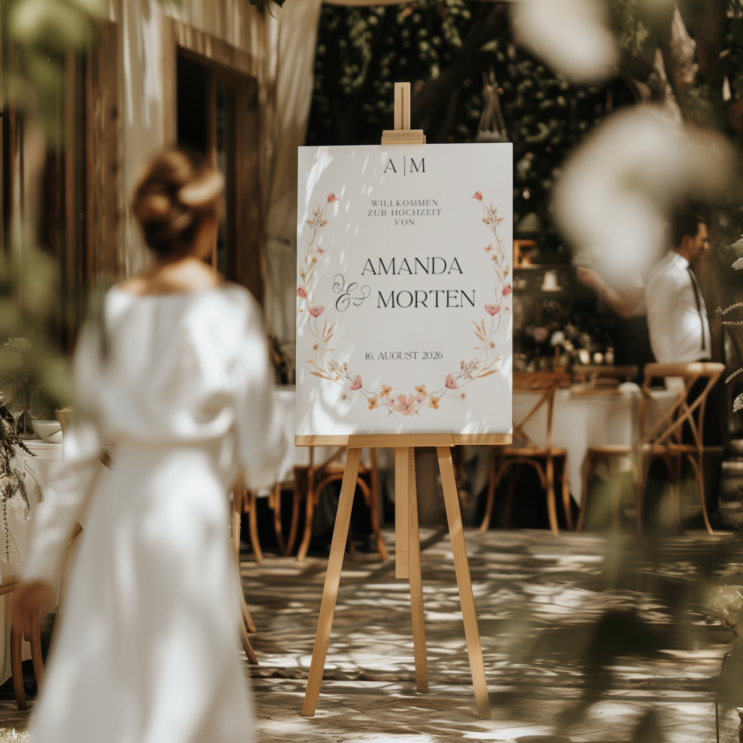 Eine Frau in einem weißen Kleid blickt auf ein elegantes Boho-Hochzeitsschild „Amanda“ von Little Tree Hill, das auf einer Staffelei ausgestellt ist. Das Willkommensschild ist wunderschön mit Blumenmustern geschmückt und zeigt die Namen „Amanda & Morten“ sowie das Datum „16. August 2026“. Der Veranstaltungsort im Freien, umgeben von Grün und Holzmöbeln, ergänzt das Boho-Hochzeitsthema perfekt.